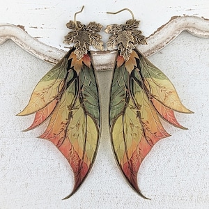 May include: A pair of dangle earrings featuring bronze-toned metal leaf-shaped toppers attached to elongated, wing-like pendants. The pendants display a gradient pattern of green, yellow, and orange hues with intricate dark brown branch and leaf vein details.