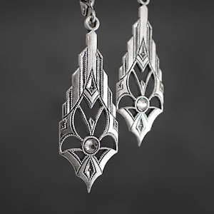 May include: A pair of silver earrings with a geometric design and clear rhinestones. The earrings are in the shape of a teardrop and have a delicate, intricate pattern.