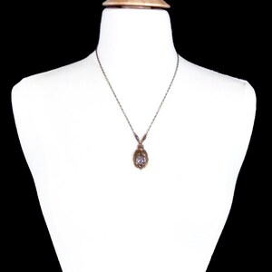 May include: A silver chain necklace with a small, oval-shaped pendant. The pendant is made of gold-toned metal and features a clear, faceted gemstone in the center.