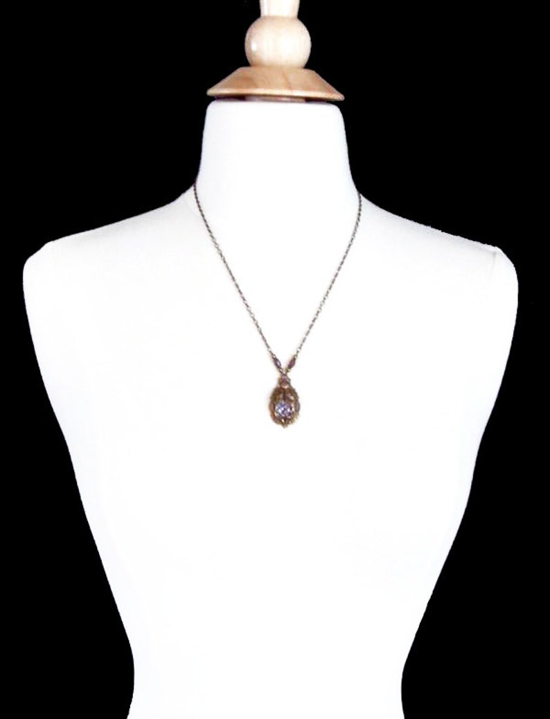 May include: A silver chain necklace with a small, oval-shaped pendant. The pendant is made of gold-toned metal and features a clear, faceted gemstone in the center.