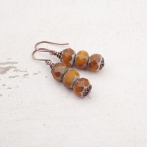 May include: A pair of handcrafted earrings with amber-coloured, faceted beads and a floral design. The beads are strung with copper-coloured metal findings, including the ear wires. The earrings are displayed on a white, textured surface.
