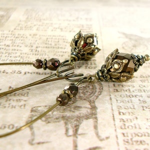 May include: A pair of gold-toned earrings with a dark brown faceted glass bead and a detailed metal design. The earrings are hanging from a thin wire.