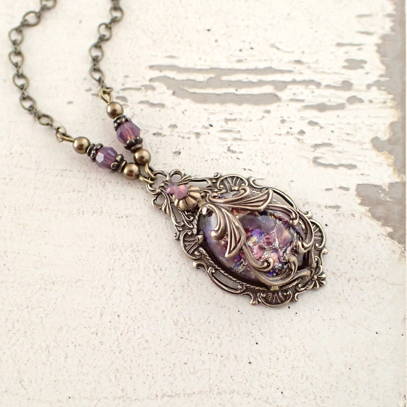 May include: A delicate necklace with a bronze chain and a pendant featuring a purple iridescent glass stone set in an ornate bronze frame.