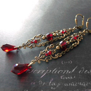 May include: A pair of long, dangling earrings with a gold filigree design and red glass beads. The earrings have a teardrop-shaped red glass pendant at the bottom.