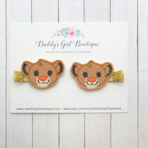 May include: Two brown lion-faced hair clips with gold-colored metal clips. The lion faces have black ears, orange noses, and embroidered white smiles. The clips are displayed on a white card with the text "Daddy's Girl Boutique".