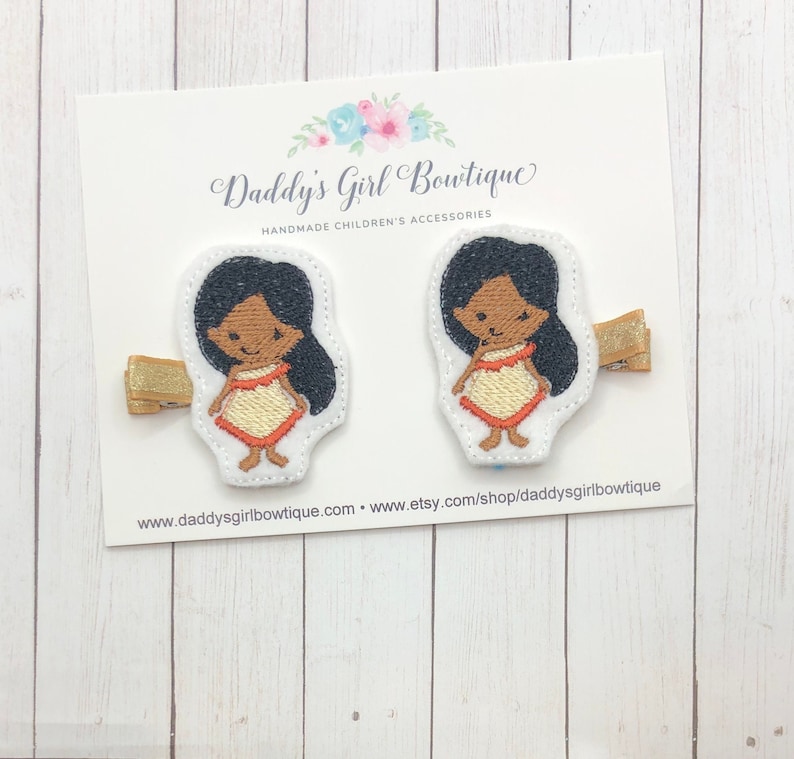 May include: Two embroidered hair clips featuring a cartoon character with black hair and a cream and orange dress. The clips are attached to gold-colored barrettes. The card reads "Daddy's Girl Boutique, Handmade Children's Accessories."
