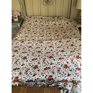 May include: A bedspread featuring a white background with a repeating pattern of red roses and green leaves. The fabric covers the entire surface of the bed, extending from the headboard down to the foot, displaying a consistent floral print throughout.