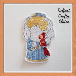 May include: An embroidered patch featuring a cartoon wizard with a long white beard, a blue robe with gold trim, and a yellow hat. The wizard holds a small red bird. The text 'Belfast Crafty Claire' is printed in black in the upper right corner.