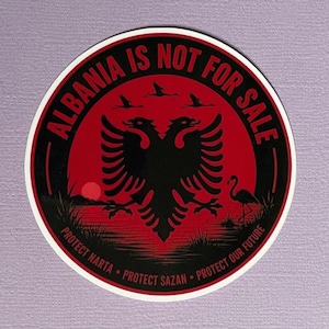 May include: A circular sticker featuring a black double-headed eagle on a red background. The text ALBANIA IS NOT FOR SALE is arched at the top. Below, the text PROTECT NARTA, PROTECT SAZAN, PROTECT OUR FUTURE is written along the bottom edge.