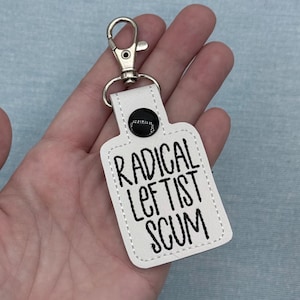 May include: White keychain with the text "RADICAL LEFTIST SCUM" in black lettering. The keychain has a silver-colored clasp and a black button. The edges are stitched with white thread.