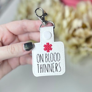 May include: White leather keychain with a red medical symbol and the text "ON BLOOD THINNERS" in black.