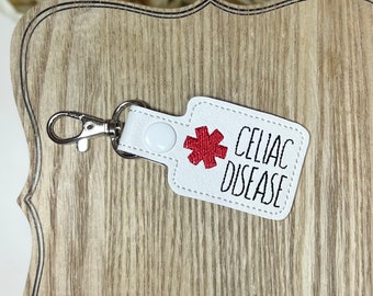 May include: White leatherette keychain with a silver clasp. The keychain has a red asterisk symbol and the words "CELIAC DISEASE" in black lettering. A white snap button secures the keychain.
