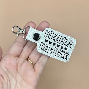 May include: White keychain with a black button and the text "Pathological People Pleaser" with five black hearts.