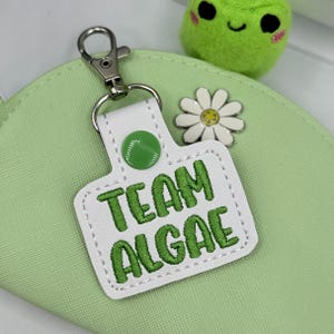 May include: A white rectangular keychain with rounded corners featuring the embroidered green text "TEAM ALGAE". The keychain has a green snap button, a silver metal lobster clasp, and is decorated with a small white daisy charm with a yellow center.