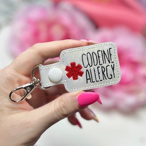 May include: A white rectangular keychain with the words "CODEINE ALLERGY" in black lettering and a red medical symbol. It features a silver clasp and a white snap closure.