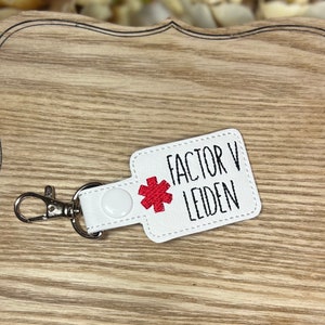May include: White keychain with the text "FACTOR V LEIDEN" and a red asterisk symbol. The keychain has a silver clasp and a white snap closure. The background is a light wood surface.