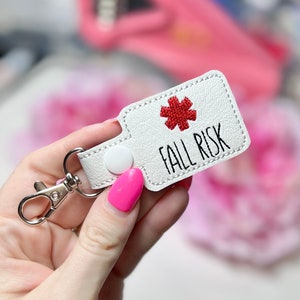 May include: White keychain with the text "FALL RISK" and a red starburst emblem. The keychain has a silver clasp and a white snap closure. The background is blurred, with pink and white elements.