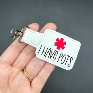May include: White leather keychain with a red medical symbol and the text "I HAVE POTS".