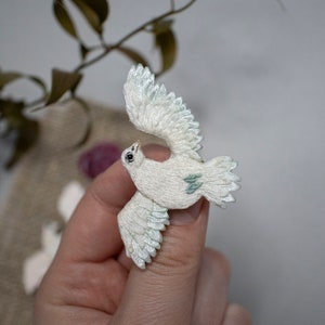 May include: A white embroidered dove brooch with green accents. The dove is depicted in flight with its wings spread.