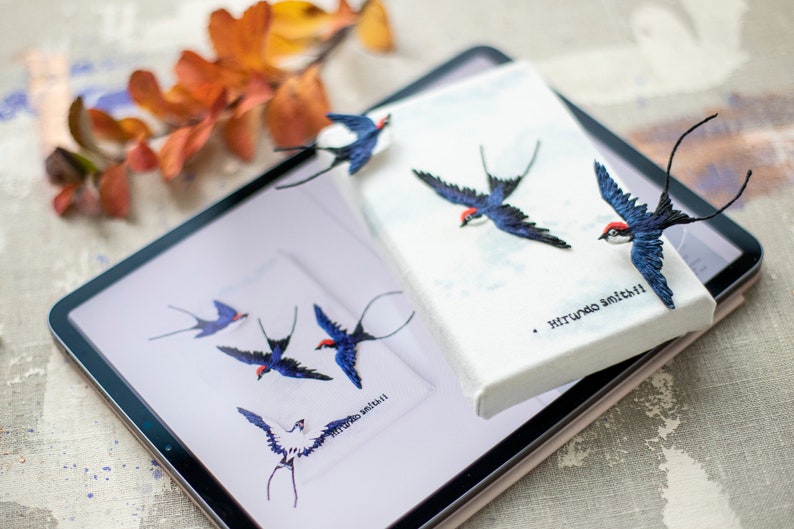 Pu&ograve; includere: Un tablet digitale che mostra immagini ricamate di rondini blu e bianche su uno sfondo bianco. Il tablet &egrave; sopra una tela bianca con una rondine ricamata blu e bianca. La tela ha il testo "Hirundo Smith" scritto su di essa.