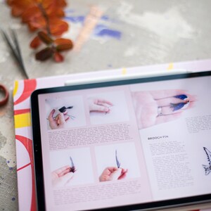 Pu&ograve; includere: Un tablet che mostra una pagina di rivista con istruzioni su come realizzare una spilla a forma di uccello. La pagina include foto dei passaggi coinvolti e una descrizione del progetto. Il testo sulla pagina recita "Brooch Pin".