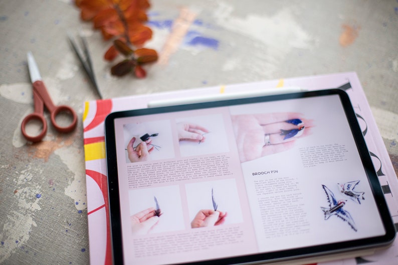 Pu&ograve; includere: Un tablet che mostra una pagina di rivista con istruzioni su come realizzare una spilla a forma di uccello. La pagina include foto dei passaggi coinvolti e una descrizione del progetto. Il testo sulla pagina recita "Brooch Pin".