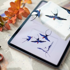 Pu&ograve; includere: Una tela bianca con rondini ricamate in blu e rosso. La tela &egrave; sopra un tablet con un'immagine simile di rondini. Il testo sulla tela recita "Mitford Mitchel".