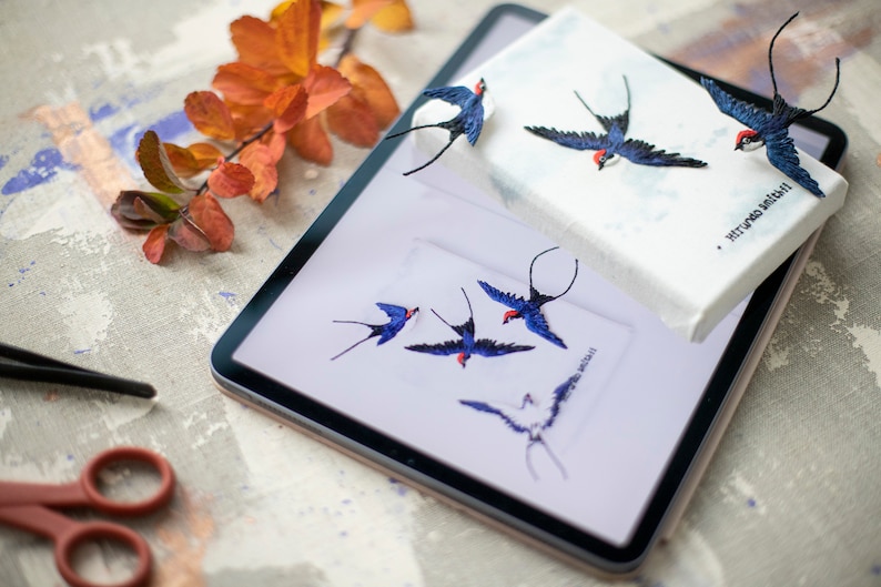 Pu&ograve; includere: Una tela bianca con rondini ricamate in blu e rosso. La tela &egrave; sopra un tablet con un'immagine simile di rondini. Il testo sulla tela recita "Mitford Mitchel".