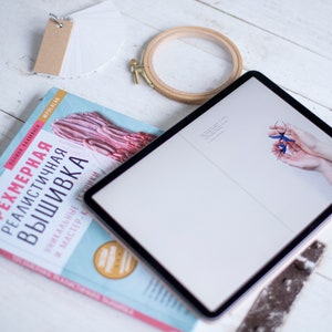 Pu&ograve; includere: Un libro blu con il titolo "Трехмерная реалистичная вышивка" su una superficie di legno bianco con un tablet, un telaio da ricamo in legno e una pila di schede di indice bianche con un'etichetta marrone.