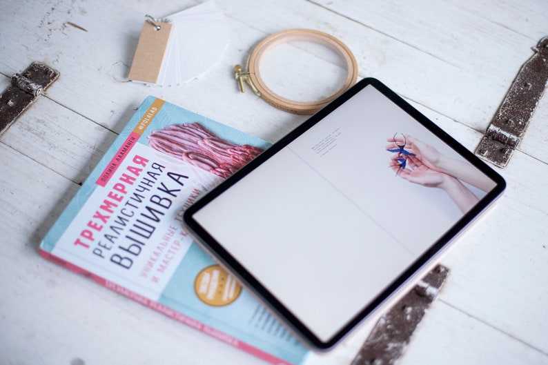Pu&ograve; includere: Un libro blu con il titolo "Трехмерная реалистичная вышивка" su una superficie di legno bianco con un tablet, un telaio da ricamo in legno e una pila di schede di indice bianche con un'etichetta marrone.