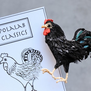 May include: A black rooster with a red comb and a teal tail feather. The rooster is made of fabric and is a handmade craft. The rooster is sitting on a white card with the text "POLALAB CLASSICS" printed on it.
