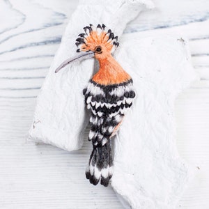 May include: A handcrafted brooch shaped like a hoopoe bird, featuring vibrant orange plumage on the head and crest, with black and white striped wings and tail feathers. The bird has a long, slender, slightly curved grey beak and a dark eye.
