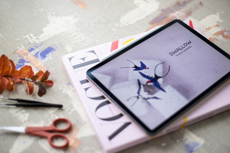 Pu&ograve; includere: Un tablet con una rivista sotto. Lo schermo del tablet mostra una copertina di rivista con il titolo "SWALLOW" e le parole "brook & street" in un carattere pi&ugrave; piccolo. La copertina della rivista presenta uno sfondo viola chiaro con un design di uccelli blu e bianchi.