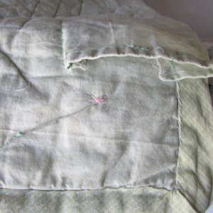 May include: Close-up of a light green quilt with a pink thread detail. The quilt is made of a soft, textured fabric and has a simple, elegant design.