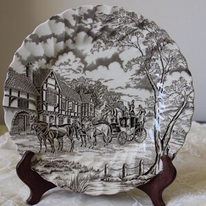 May include: A decorative plate with a scalloped edge, featuring a black and white transferware design. The scene depicts a horse-drawn carriage in front of a building, with trees and a cloudy sky. The plate is displayed on a wooden stand.