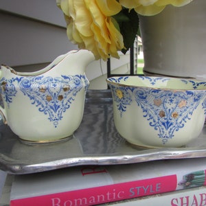 May include: A vintage cream-colored creamer and sugar bowl set with blue and gold floral designs. The set is on a silver tray and a stack of books with the text "Romantic Style" and "Shabby Chic".