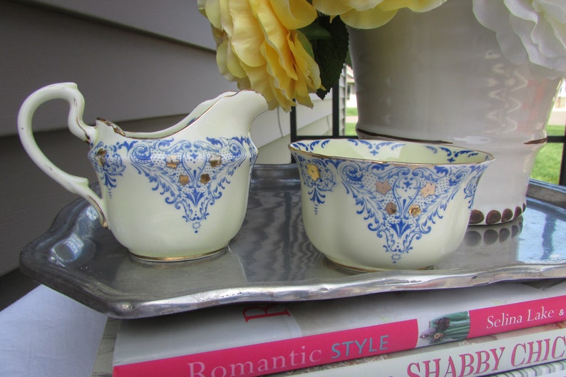 May include: A vintage cream-colored creamer and sugar bowl set with blue and gold floral designs. The set is on a silver tray and a stack of books with the text "Romantic Style" and "Shabby Chic".