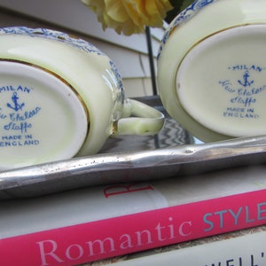 May include: A vintage cream-colored creamer and sugar set with blue and gold accents. The bottom of each piece features a blue and white logo that reads "Milan New Chelsea Staffs Made in England".