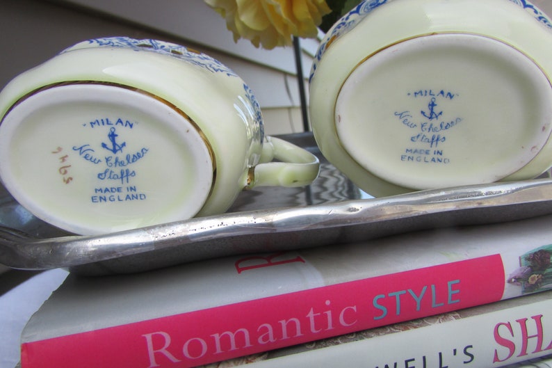May include: A vintage cream-colored creamer and sugar set with blue and gold accents. The bottom of each piece features a blue and white logo that reads "Milan New Chelsea Staffs Made in England".