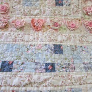 May include: A white and blue patchwork quilt with a floral pattern. The quilt is decorated with pink fabric flowers and buttons.