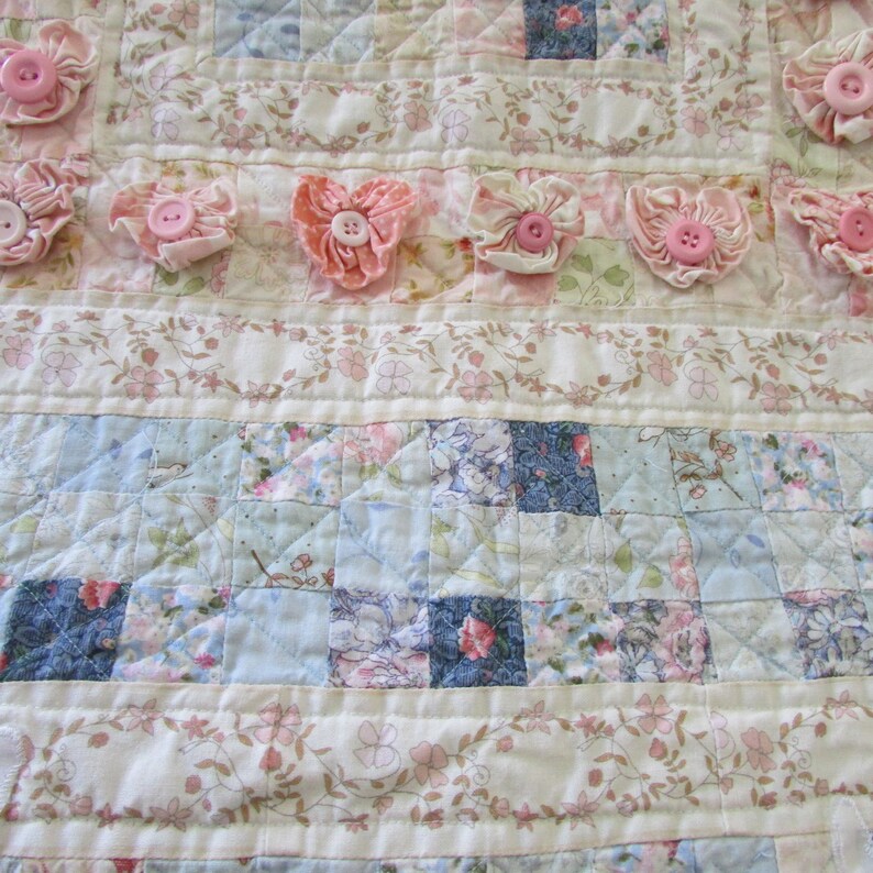 May include: A white and blue patchwork quilt with a floral pattern. The quilt is decorated with pink fabric flowers and buttons.