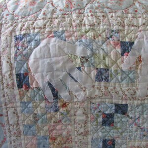 May include: A white quilted fabric with a floral pattern. Two white fabric hands are sewn onto the fabric. The hands are positioned to look like they are holding something.