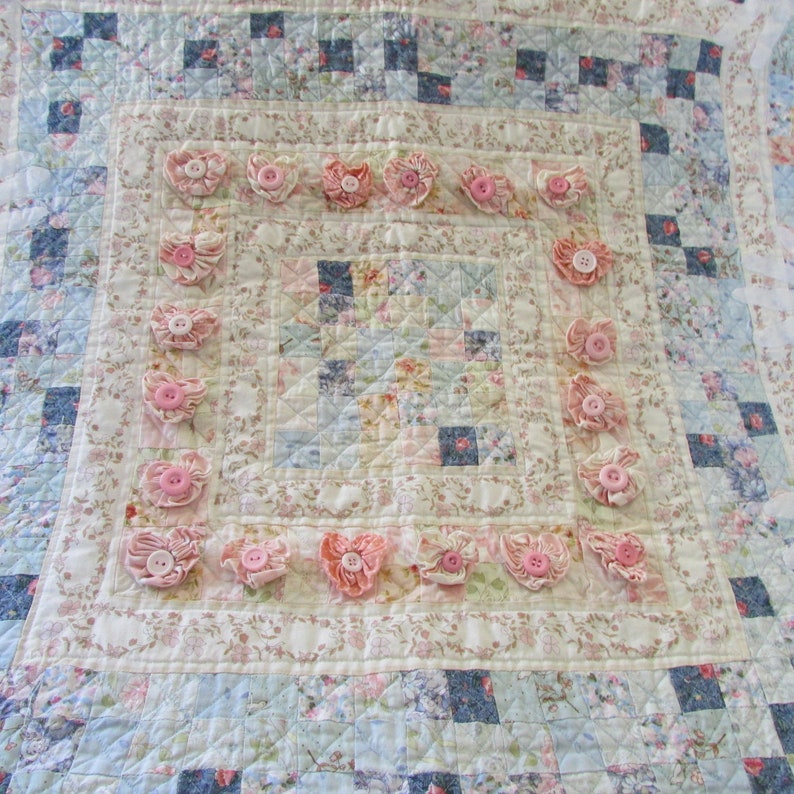 May include: A white and blue patchwork quilt with a pink floral design. The quilt is decorated with pink fabric flowers around the edges.