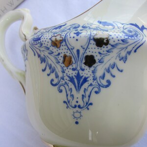 May include: A white ceramic creamer with a blue and gold floral design. The creamer has a handle and a spout.