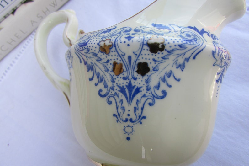 May include: A white ceramic creamer with a blue and gold floral design. The creamer has a handle and a spout.