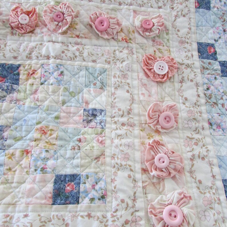 May include: A white quilt with a patchwork design featuring floral patterns in shades of pink, blue, and green. The quilt is decorated with pink fabric flowers and buttons.