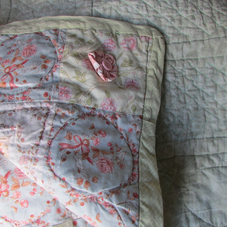 May include: A patchwork quilt with a floral pattern in shades of pink, blue, and white. The quilt is made of different fabrics and has a quilted texture. A small pink button is sewn onto the quilt.