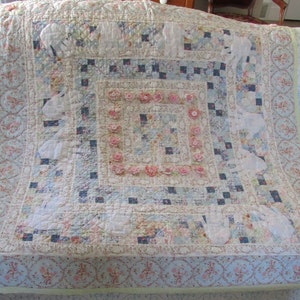 May include: A white and blue patchwork quilt with a floral border and a central square design. The quilt is decorated with small pink flowers and white appliques.