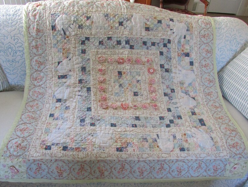 May include: A white and blue patchwork quilt with a floral border and a central square design. The quilt is decorated with small pink flowers and white appliques.
