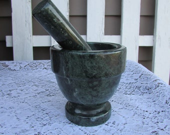 May include: A green marble mortar and pestle set. The mortar is a deep bowl with a wide base and a slightly flared rim. The pestle is a long, cylindrical shape with a rounded end.